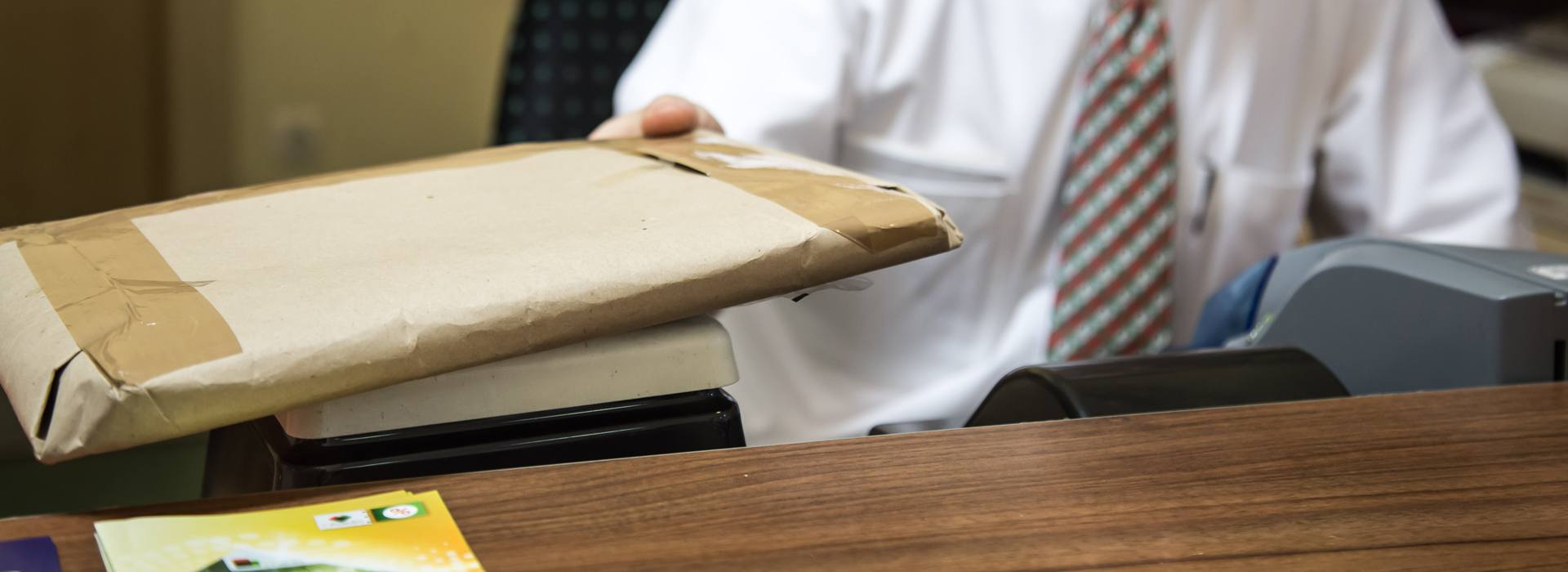 A képen egy nyakkendős ügyintéző látható, aki egy lapos csomagot mér le egy kis postai mérlegen.