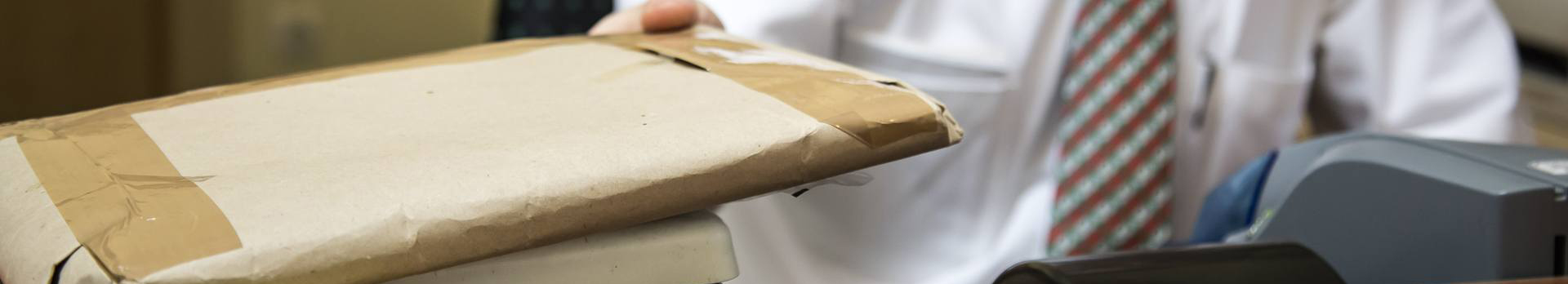 A képen egy nyakkendős ügyintéző látható, aki egy lapos csomagot mér le egy kis postai mérlegen.